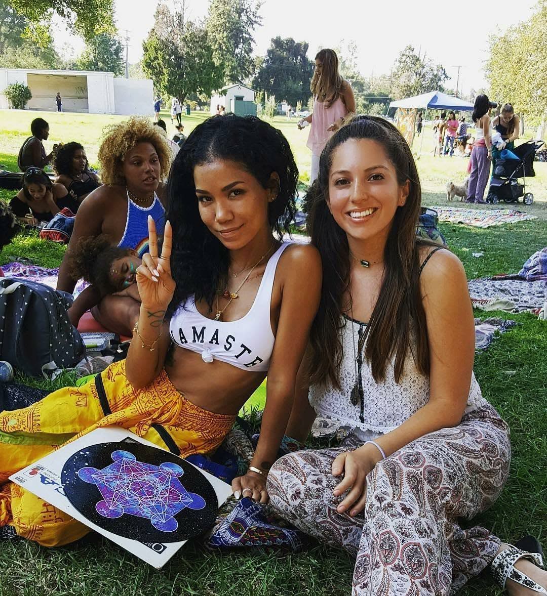 Two smiling friends on the grass amid the crowd, one in a Namaste top holding a Metatron's-cube print.