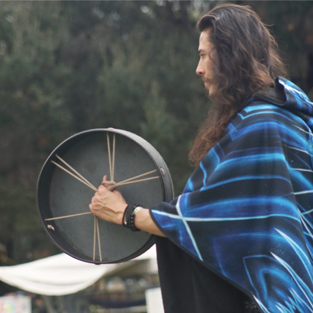 A long-haired musician in a glowing electric-blue patterned blanket holding a hand frame-drum against dark foliage.