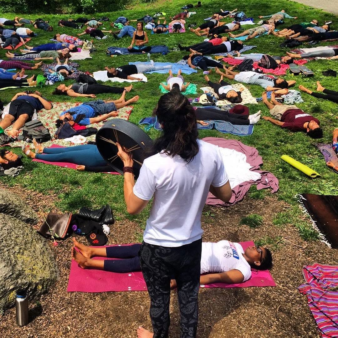 A medicine drummer walking among dozens of people lying in savasana on the grass.