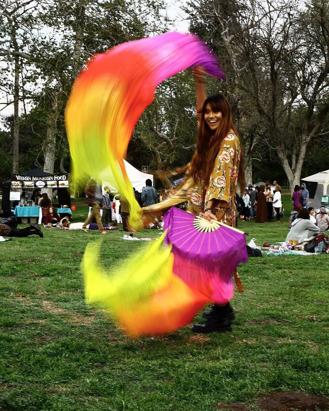 A dancer spinning vivid rainbow silk fans in motion-blur on the green park grass, the soul-family picnic behind her.