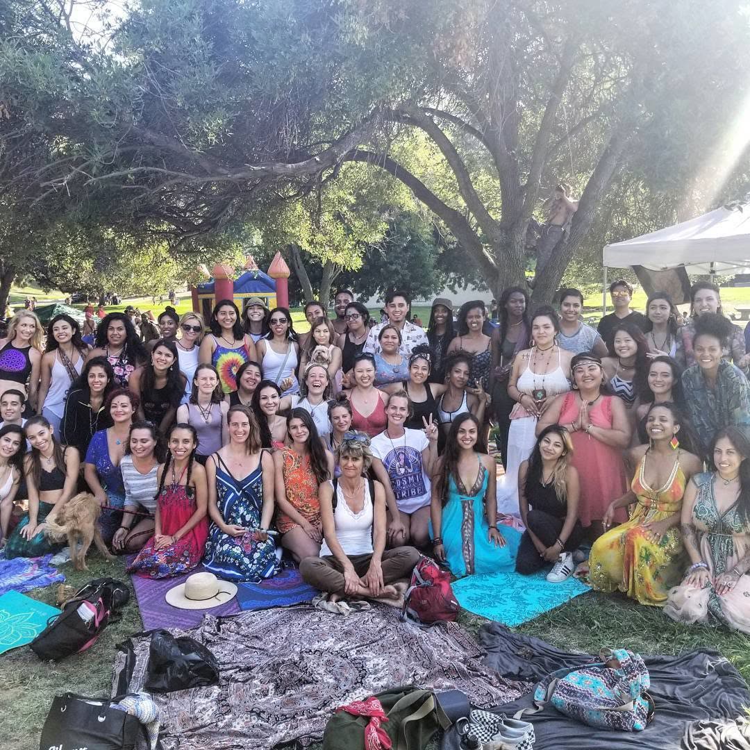 A joyful large group of women, many in flowing dresses, gathered under a big oak tree for a Divine Feminine circle.