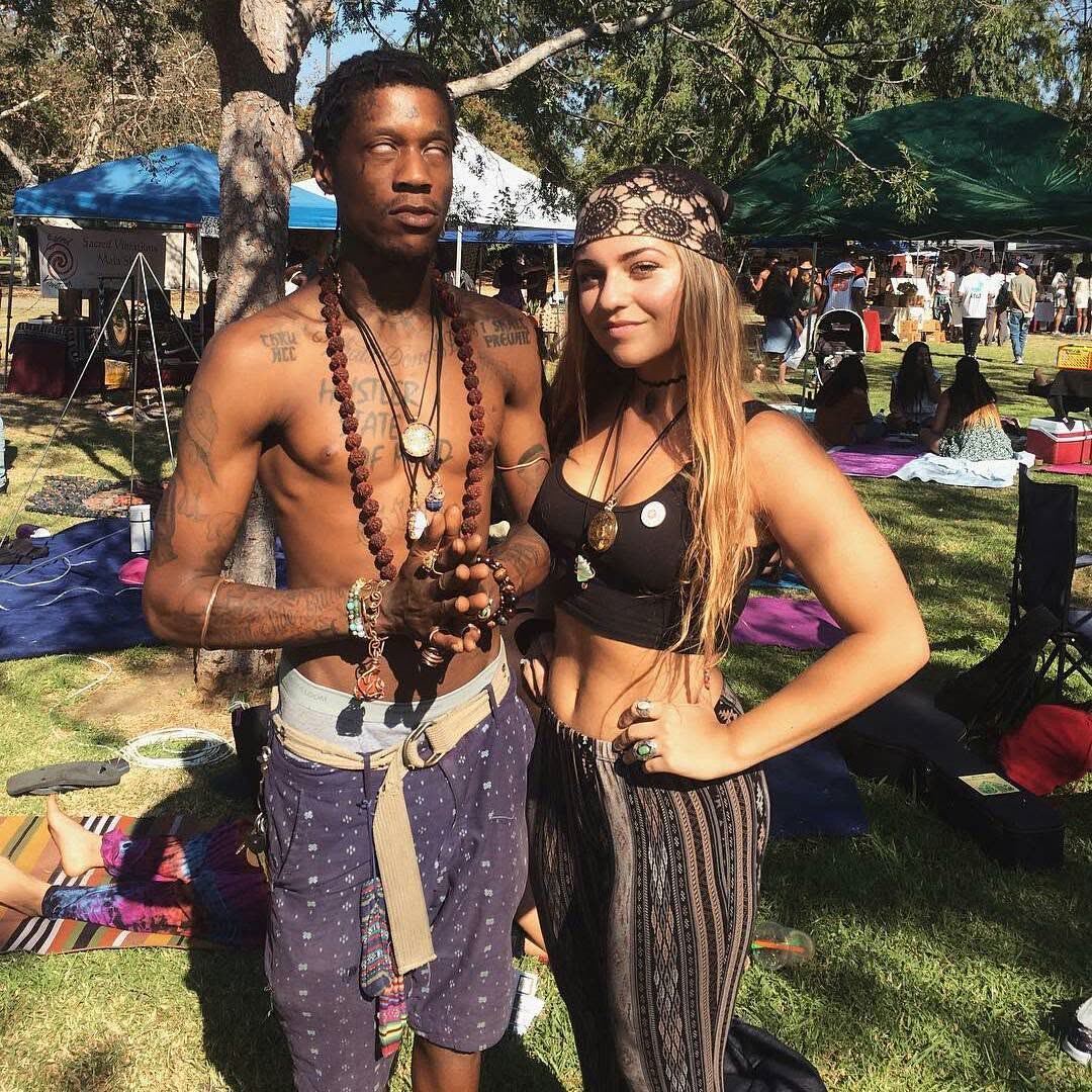 Conscious hip-hop artist draped in mala beads standing with a friend amid the blanket-strewn park crowd.