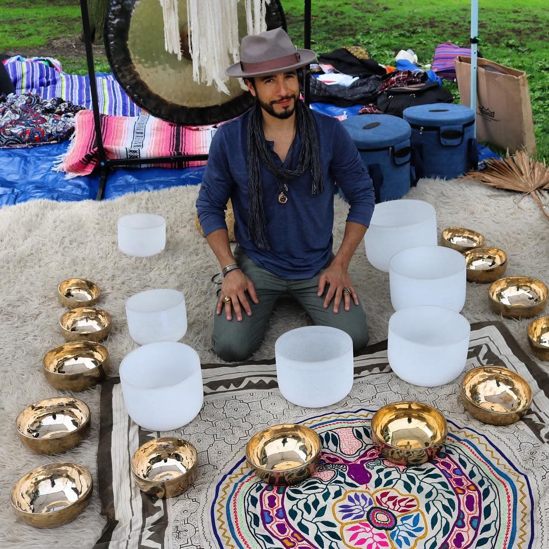 A sound healer kneeling inside a full circle of crystal singing bowls around a painted floral mandala, with a gong behind.