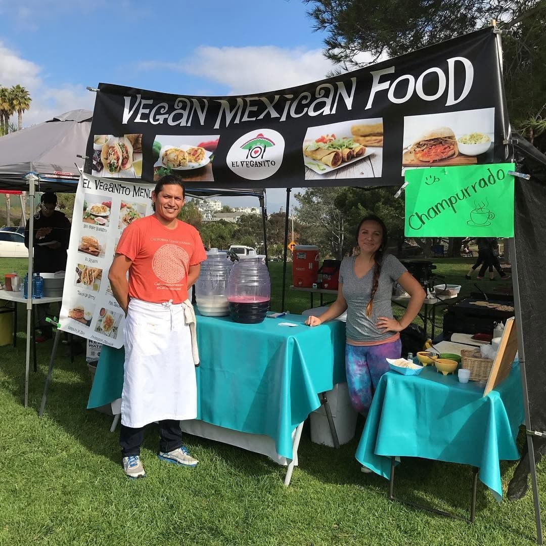 A 'Vegan Mexican Food' booth with two smiling vendors, teal tablecloths and hand-lettered menu signs on the park grass.