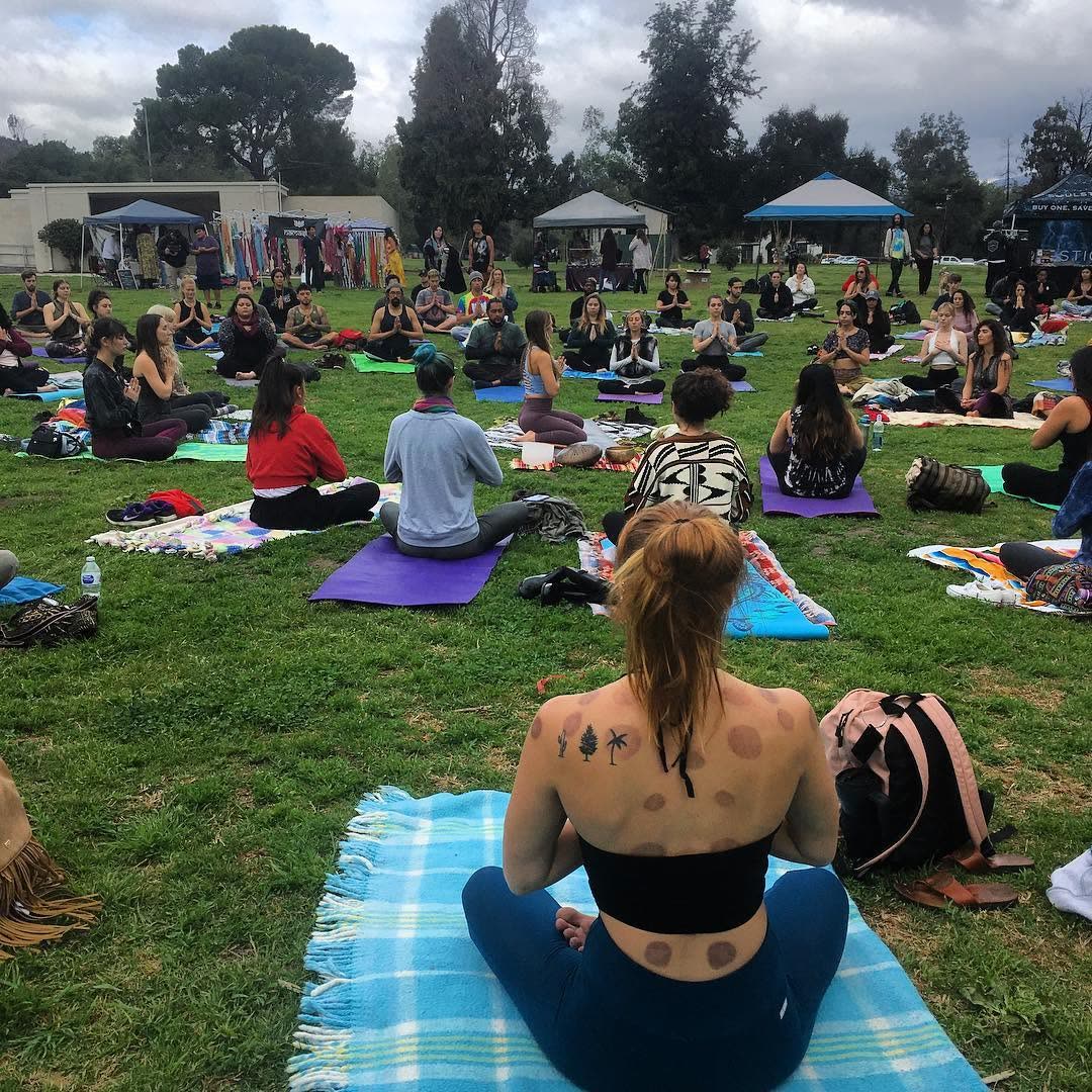 Dozens of people seated on yoga mats and blankets on the grass under a cloudy Los Angeles sky during a community yoga flow.